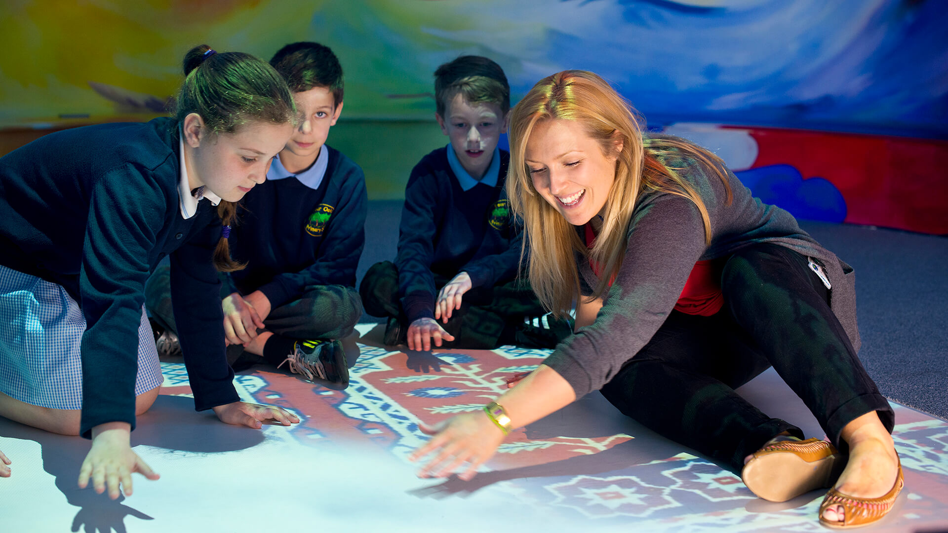A female teacher sits on the floor with three primary aged students. It is an interactive floor and they are learning from it. 