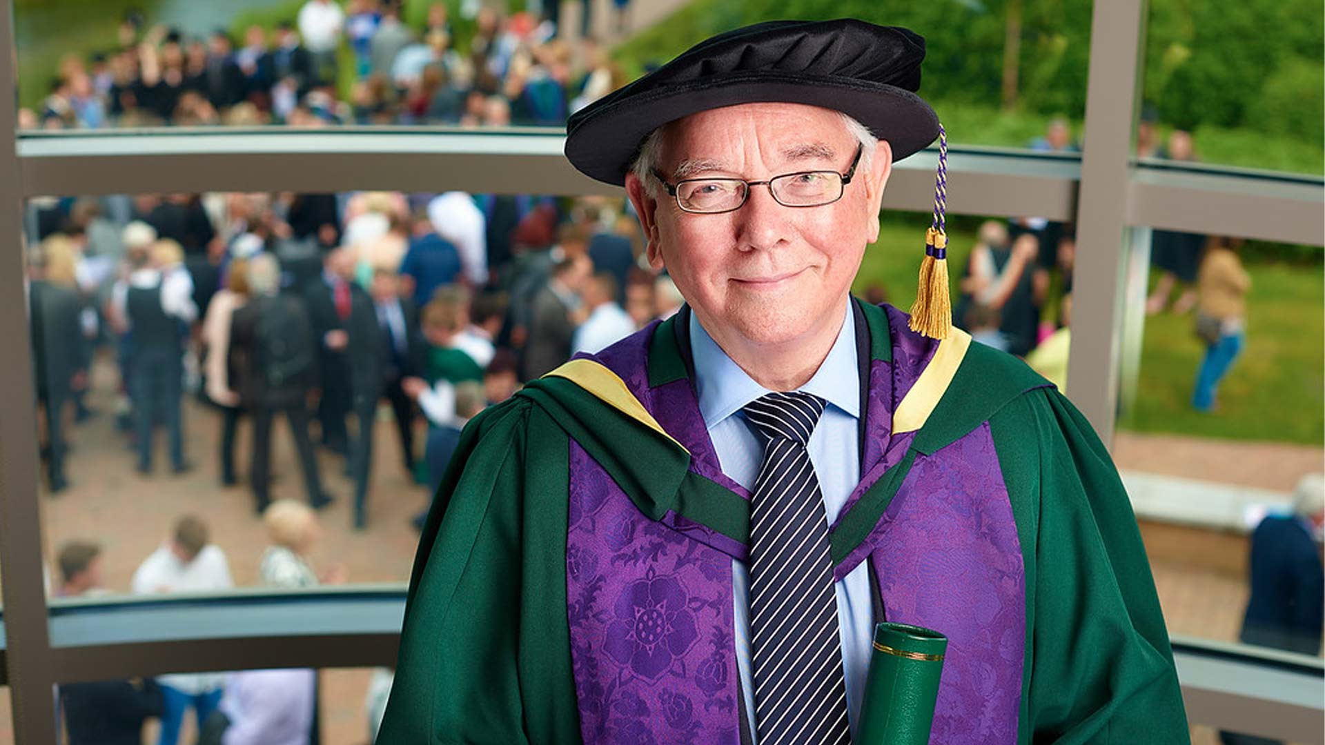 Terence Davies wears a cap and gown graduates and their supporters are seen celebrating in the background.