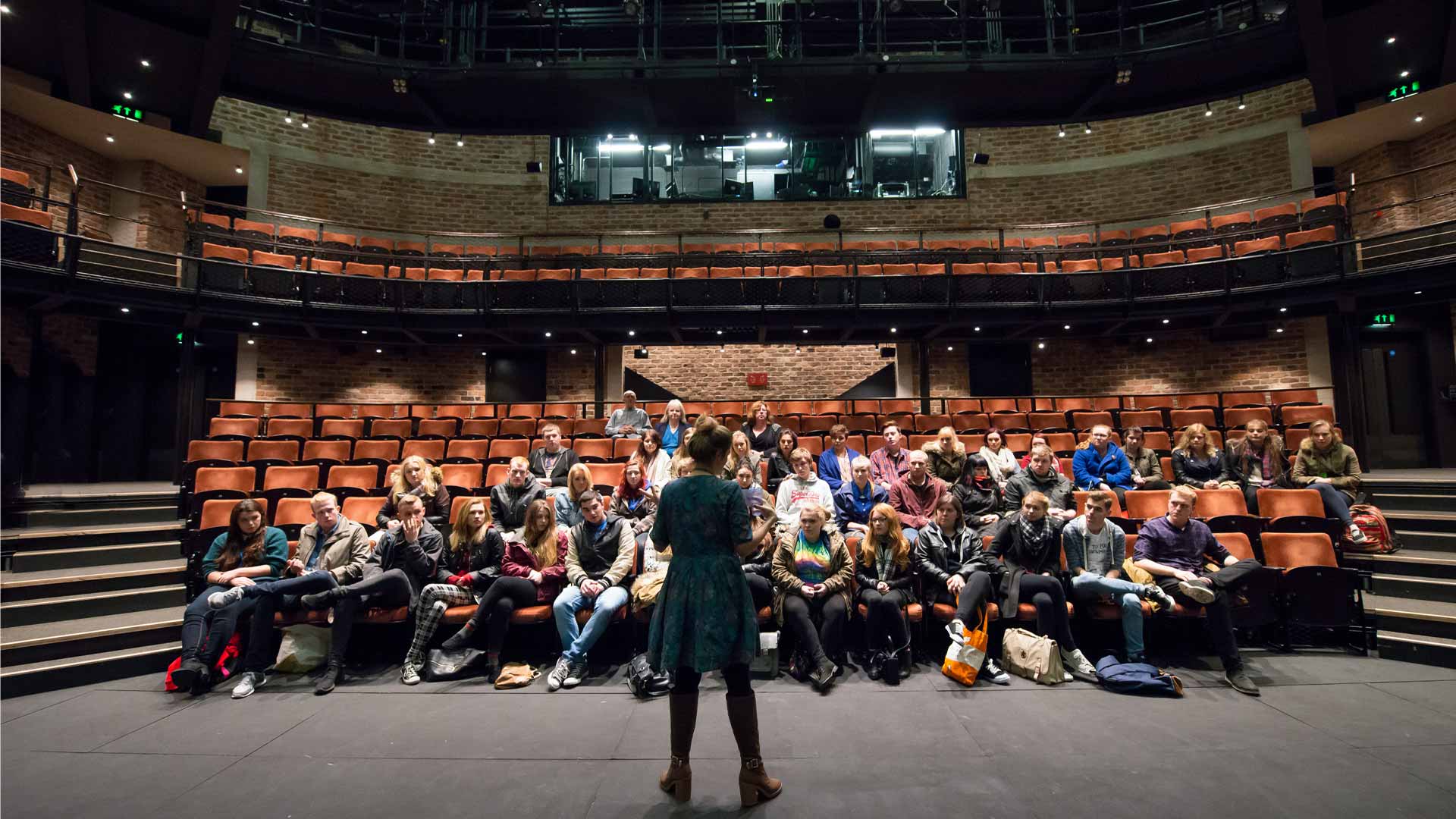 A lady stands at the front of a theatre, addressing a group of people who are sat in the audience. 