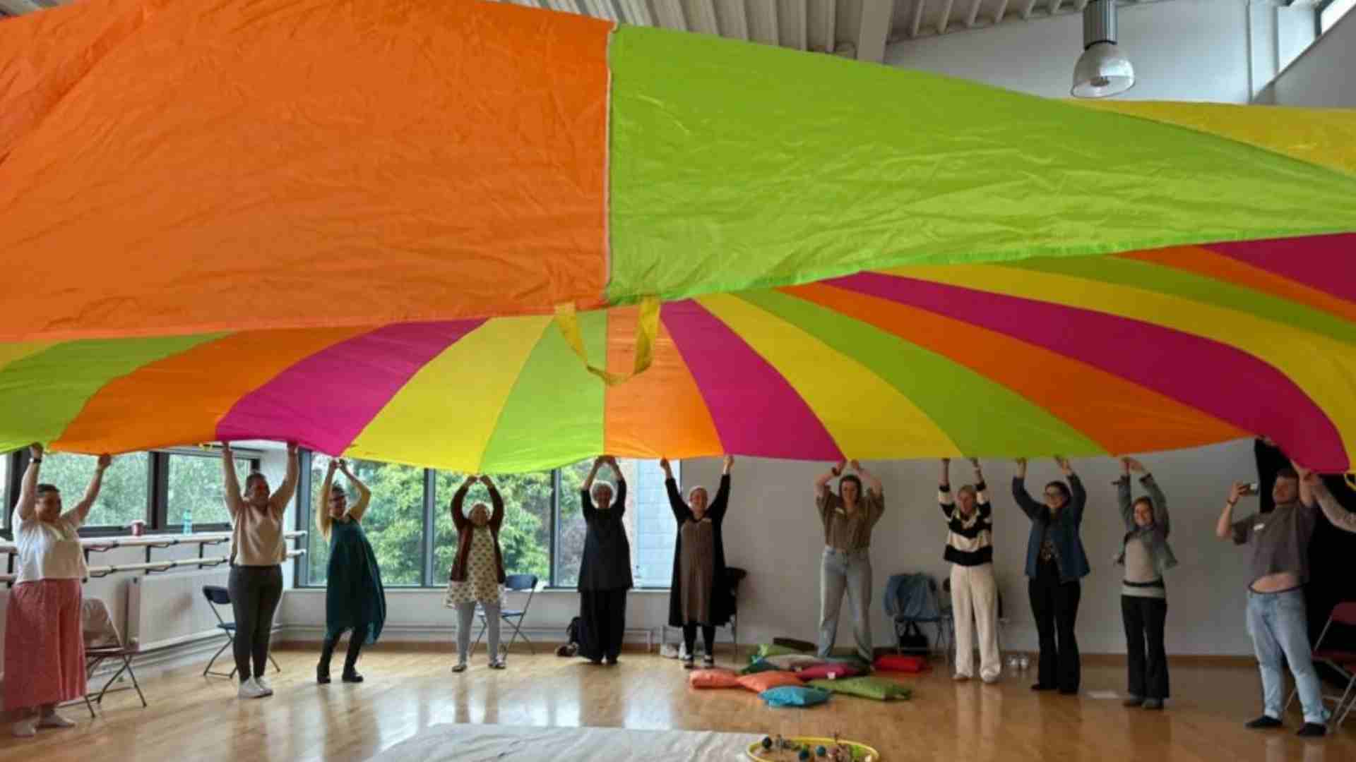 A group of adults attending Arts for the Blues training lift up a colourful parachute.